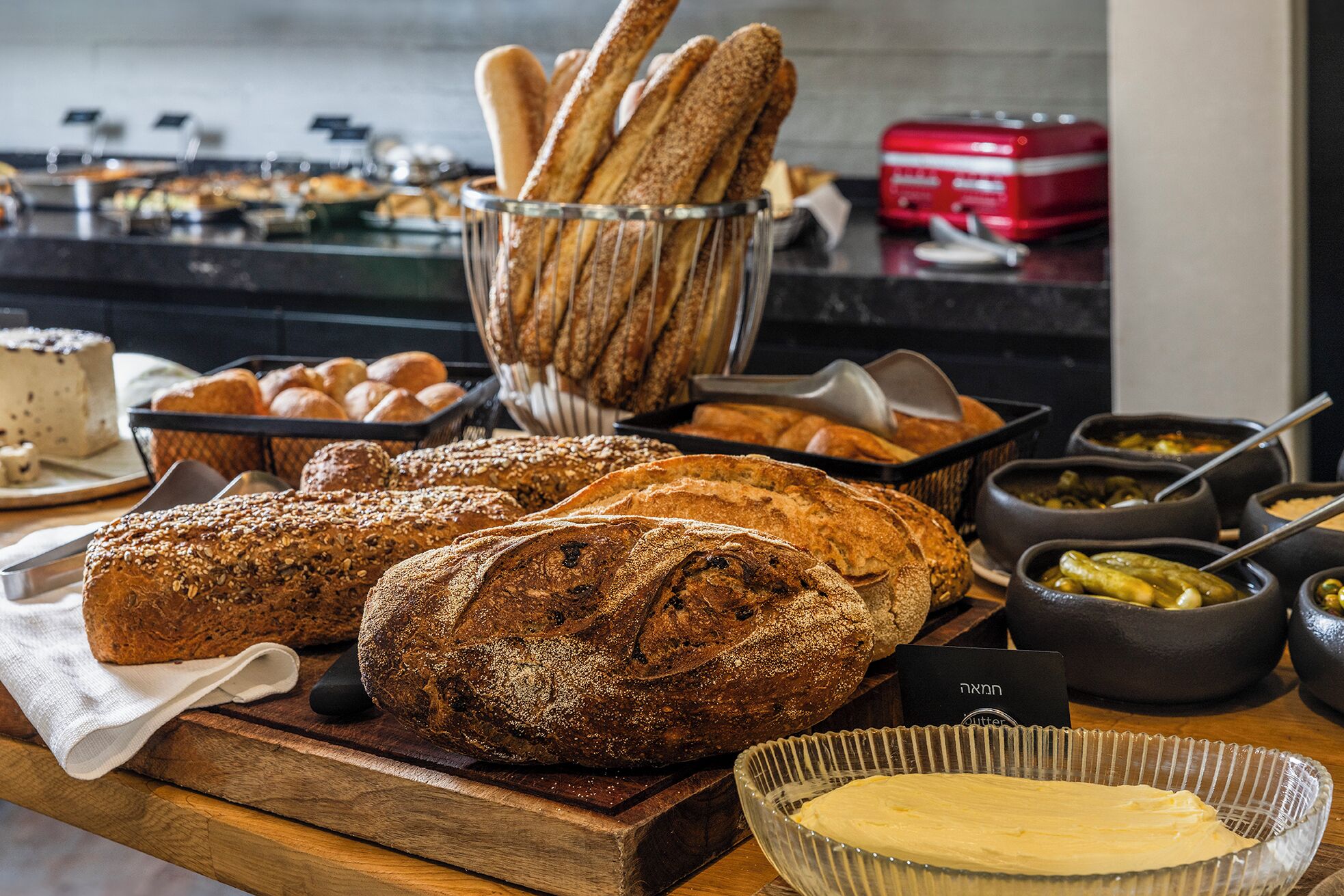 Verschiedene Brotsorten und Gebäck auf einem Holzbrett, mit einer Butter-Schale und einem roten Gerät im Hintergrund.