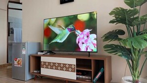 Una sala de estar con un televisor de pantalla plana sobre un soporte de madera, un refrigerador, una planta en maceta y un cuadro colgado en la pared.
