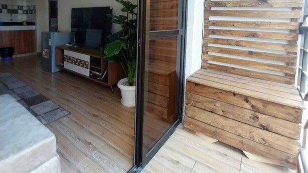 Un salón moderno con un ventanal corredera que da a un balcón, un televisor, una planta y una cocina al fondo.