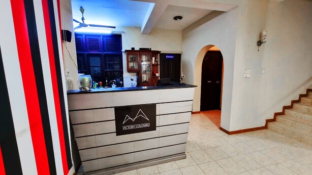 Un espace d’accueil avec un comptoir, une enseigne indiquant « Victory Colombo » et un escalier menant à une porte portant l’inscription « Cuisine ».