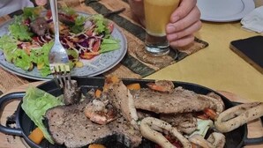 Une table à manger avec une assiette de viande et de légumes grillés, une fourchette et un verre contenant une boisson.