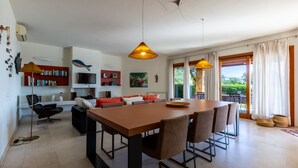 Un amplio salón con una gran mesa de comedor de madera, asientos cómodos, una estantería, un televisor y vistas al exterior a través de grandes ventanales.