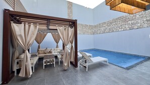 Zona de piscina con área de estar techada, piscina y pared de piedra.