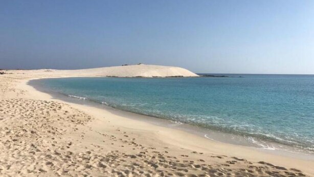 Una playa arenosa con agua azul cristalina y una duna de arena blanca al fondo.