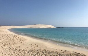 En sandstrand med krystalklart blåt vand og en hvid sandklit i baggrunden.