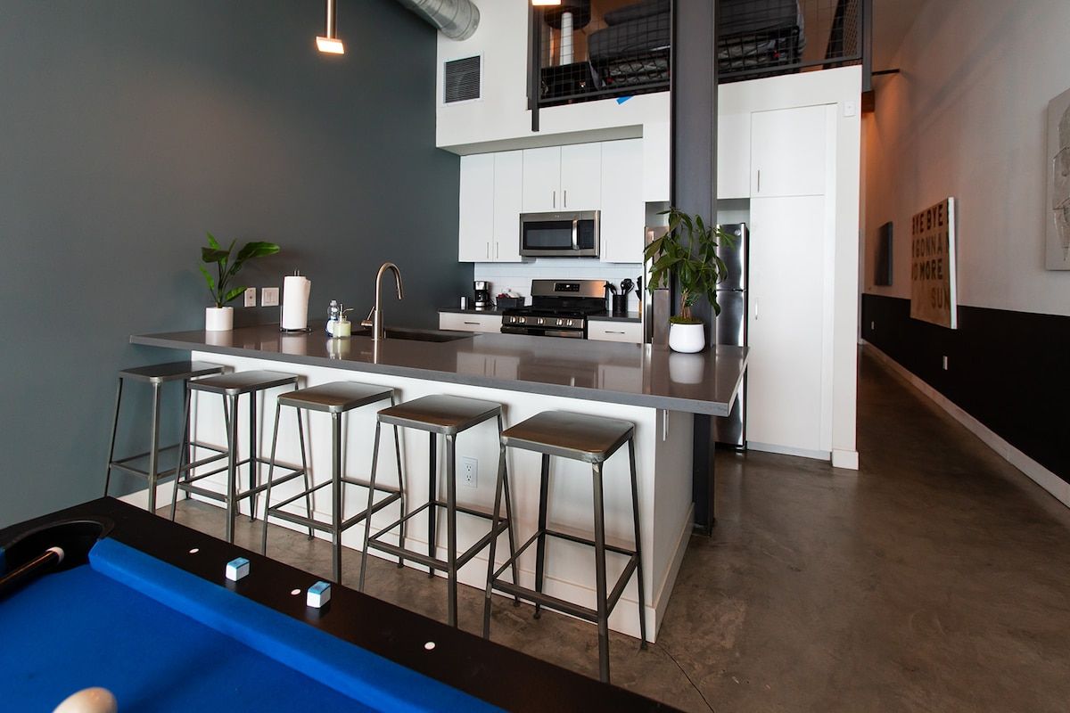 A modern kitchen with a central island, bar stools, and a pool table in the foreground.