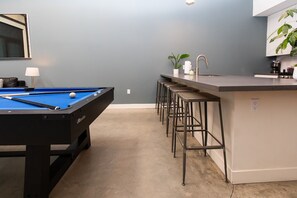 Une table de billard recouverte d’un drap bleu, un coin bar avec des tabourets et un comptoir de cuisine avec un évier et des plantes.
