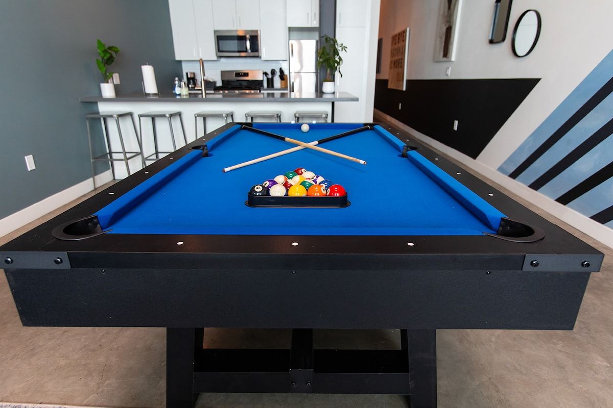 A pool table with a set of balls and cues, a kitchen area with bar stools, and a microwave.