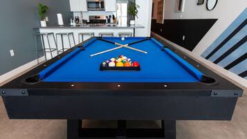 A pool table with a set of balls and cues, a kitchen area with bar stools, and a microwave.