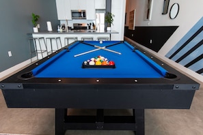 A pool table with a set of balls and cues, a kitchen area with bar stools, and a microwave.