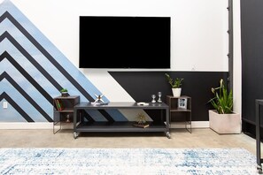 A modern living room with a flat-screen TV mounted on the wall, a black entertainment unit with decorative items, and a blue and white patterned rug. - 5Mile to Disney • Gym • Pool Table • Pet Ok • Parking (Santa Ana)