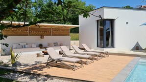 Un espace piscine extérieur moderne, doté de chaises longues, d’une terrasse en bois et d’un bâtiment blanc avec de grandes fenêtres.