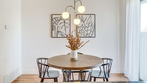 A dining area with a round wooden table, four chairs, a vase with dried flowers, and two framed artworks on the wall.