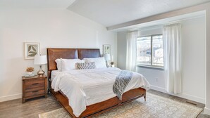 A bedroom with a wooden bed, white bedding, a nightstand with a lamp, and a window with curtains.