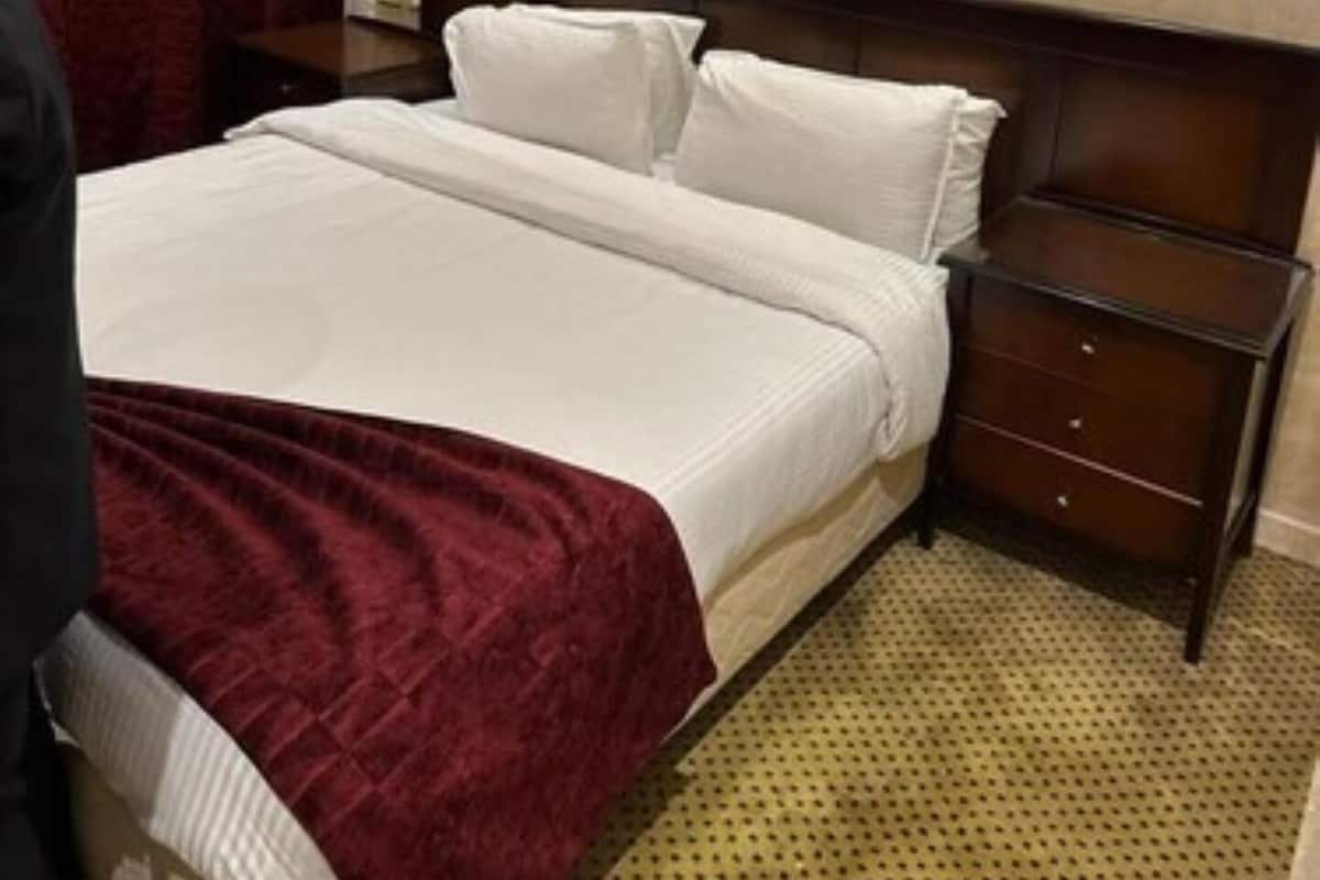 A hotel room with a large bed, a wooden headboard, a nightstand with drawers, a painting on the wall, and a red curtain.