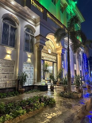 L’entrée d’un hôtel, sur laquelle on peut lire l’inscription « Alfouad Palace Hotel & Spa », se distingue par une grande arche, des panneaux décoratifs sur les murs et un chemin bien éclairé, agrémenté de plantes en pot.