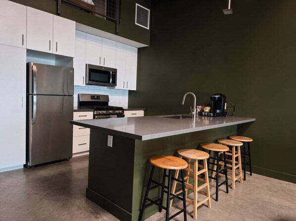 A modern kitchen with a stainless steel refrigerator, a microwave, a stove, and a coffee maker. There are four wooden stools at the island counter.