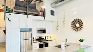Een moderne keuken met witte kastjes, roestvrijstalen apparatuur, een centraal eiland met krukken en een slaapkamer op een entresol.