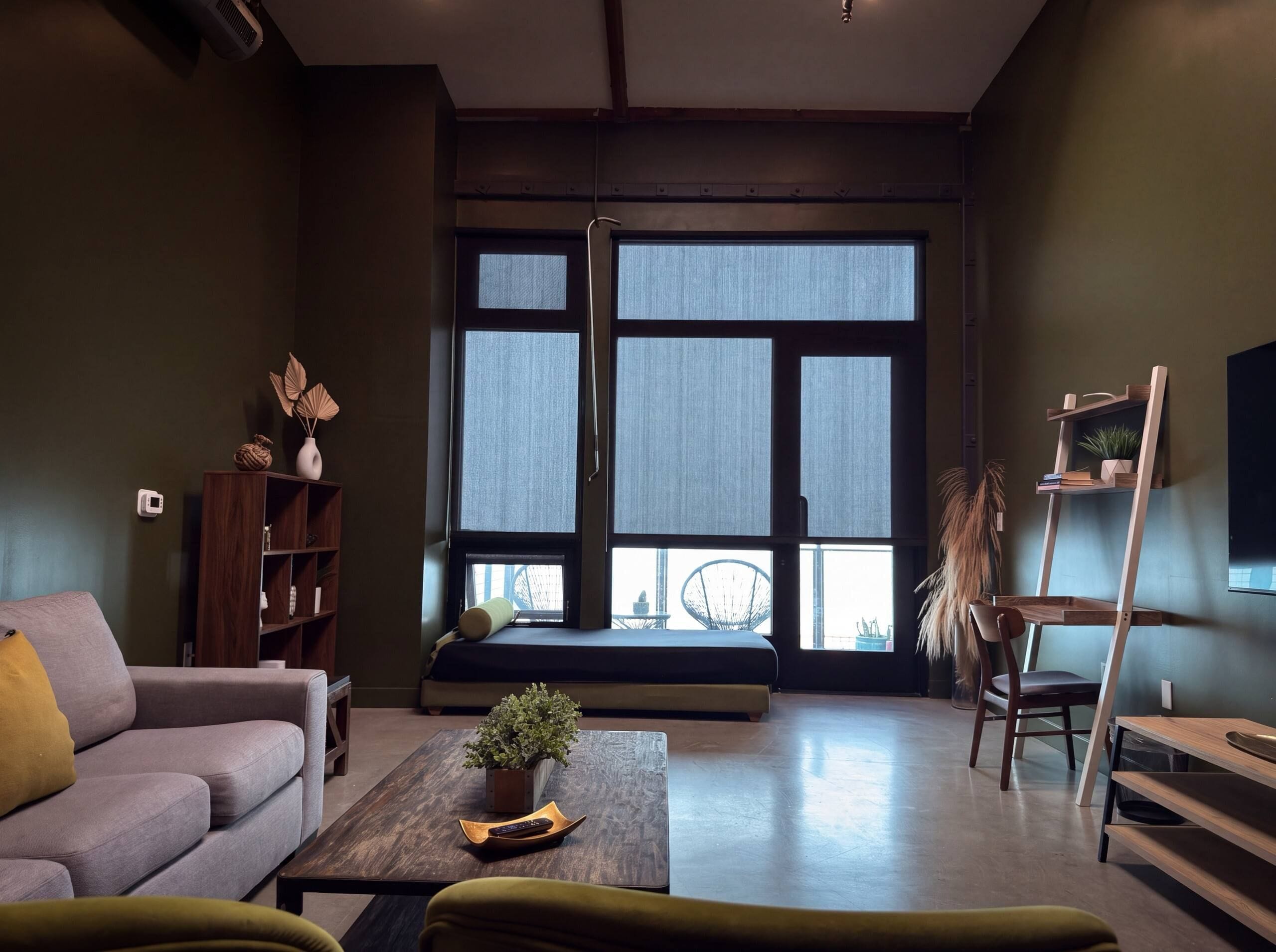 A living room with a sofa, a coffee table with a plant, a bookshelf, and a large window with blinds.
