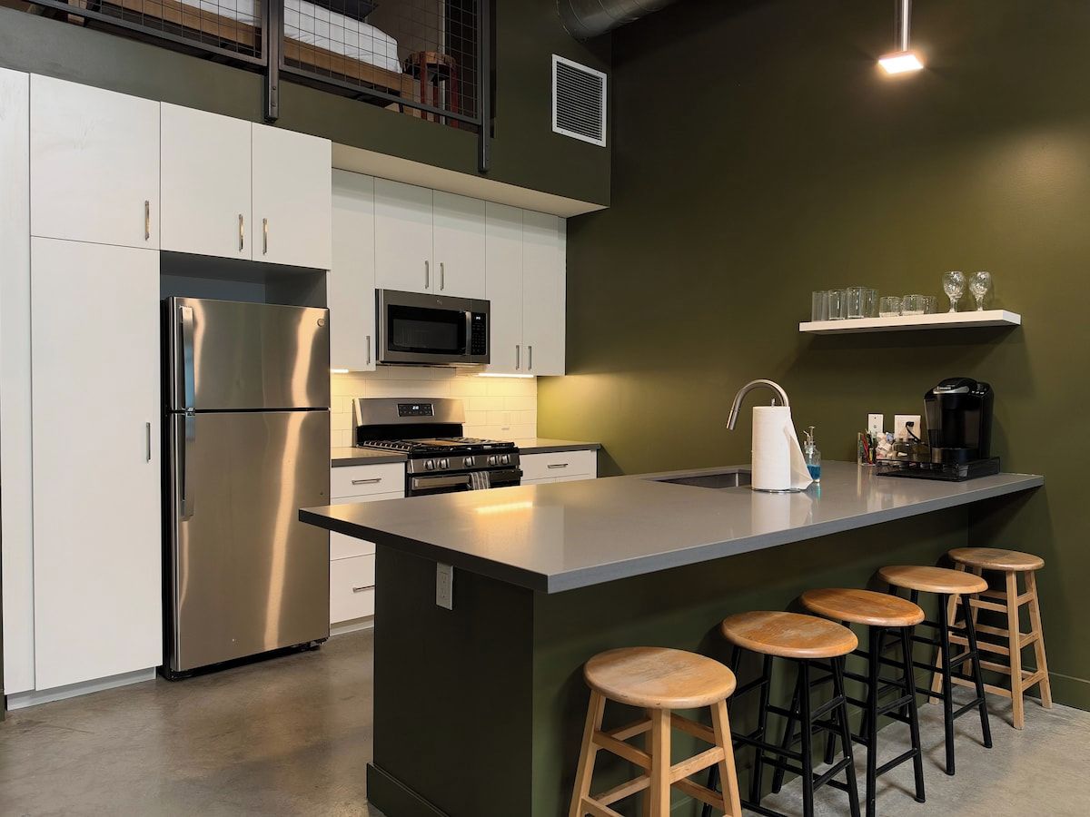 A modern kitchen with a stainless steel refrigerator, a microwave, a stove, and a coffee maker. There are four wooden stools around the bar area.
