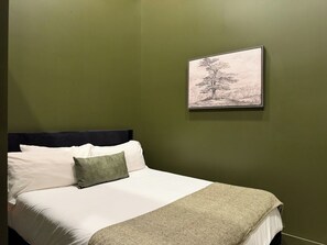 Een bed met witte lakens en een groen kussen, een grijze plaid en een lijst met een foto van een boom aan een groene muur.