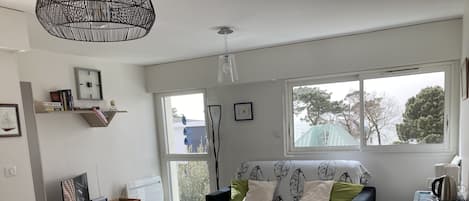 A modern living room with a sofa, a dining table, and chairs. There is a window with a view of trees and buildings.
