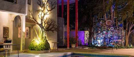A swimming pool with a tree sculpture, red pillars, and a colorful wall art installation.