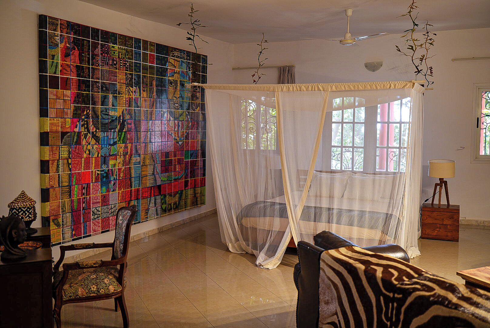 A room with a large colorful mosaic wall, a canopy bed, a chair, and a lamp.