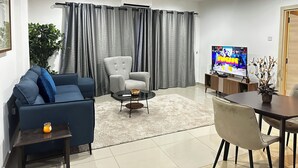 Ein modernes Wohnzimmer mit einem blauen Sofa, einem grauen Sessel, einem Couchtisch, einem Fernseher und einem Essbereich mit Stühlen und Tisch.