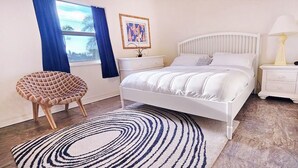 A bedroom with a bed, a bedside table with a lamp, a chair, a rug, a dresser, and a window with curtains. - The Everglades House (Ochopee)