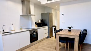 Une cuisine moderne avec des meubles blancs, un four encastré, un réfrigérateur en acier inoxydable et un coin repas comprenant une table et des chaises en bois.