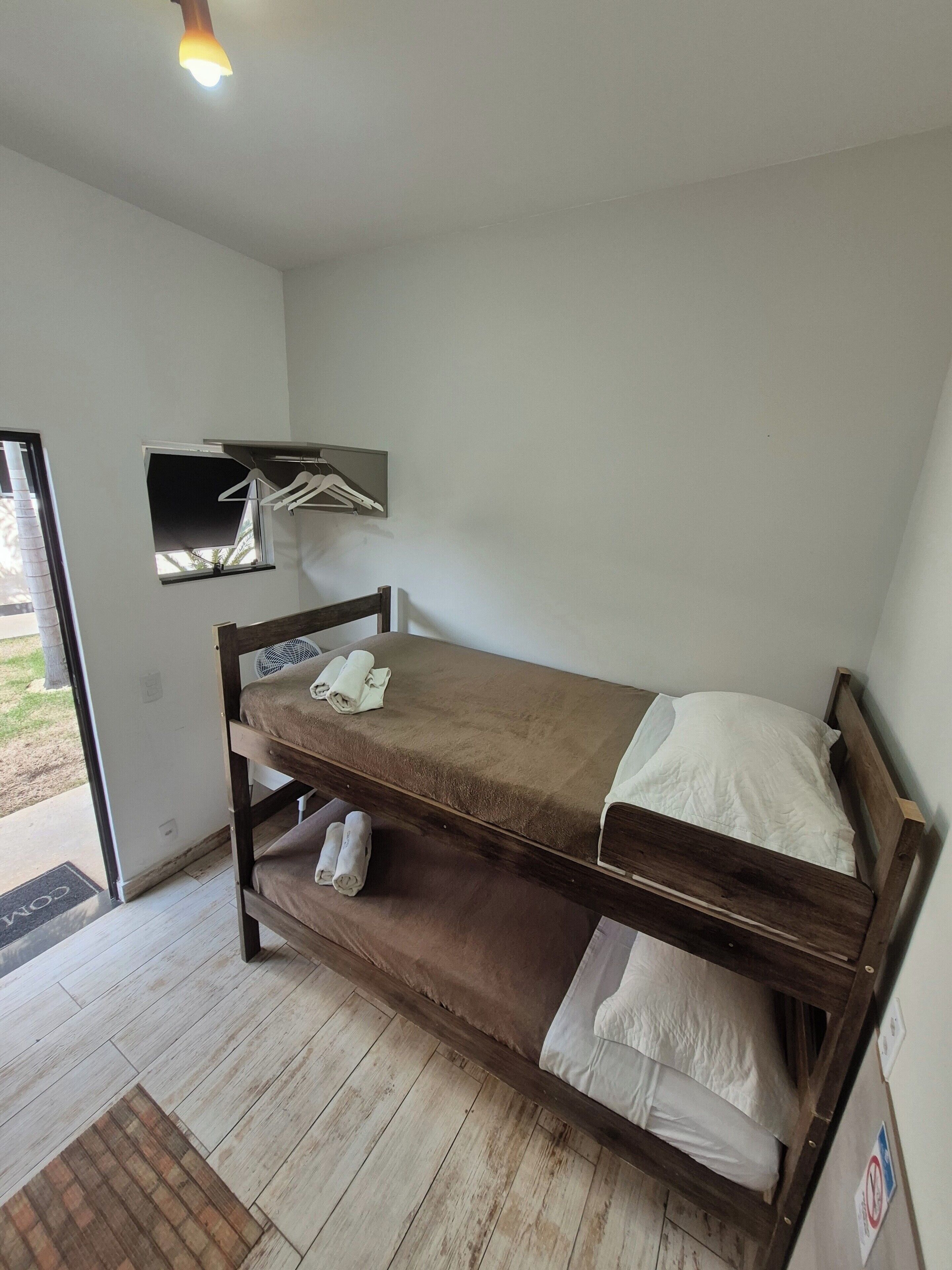 Una cama individual sobre otra con estructura de madera y un colchón marrón. Hay un estante con un ventilador y un perchero de pared con algunos ganchos. El cuarto tiene piso de madera y una puerta que da al exterior.