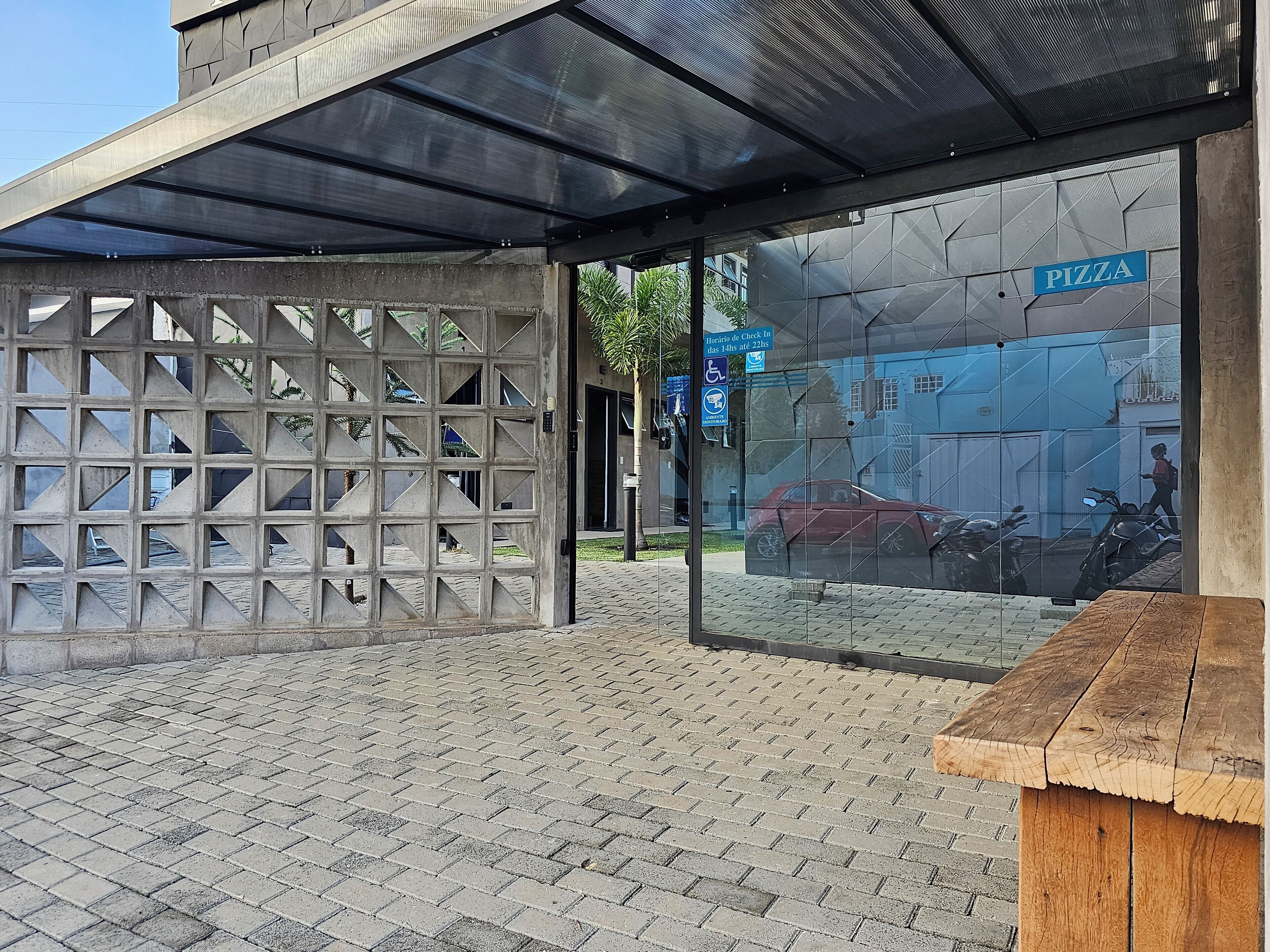 A covered outdoor seating area with a glass partition, a wooden bench, and a pizza shop in the background.