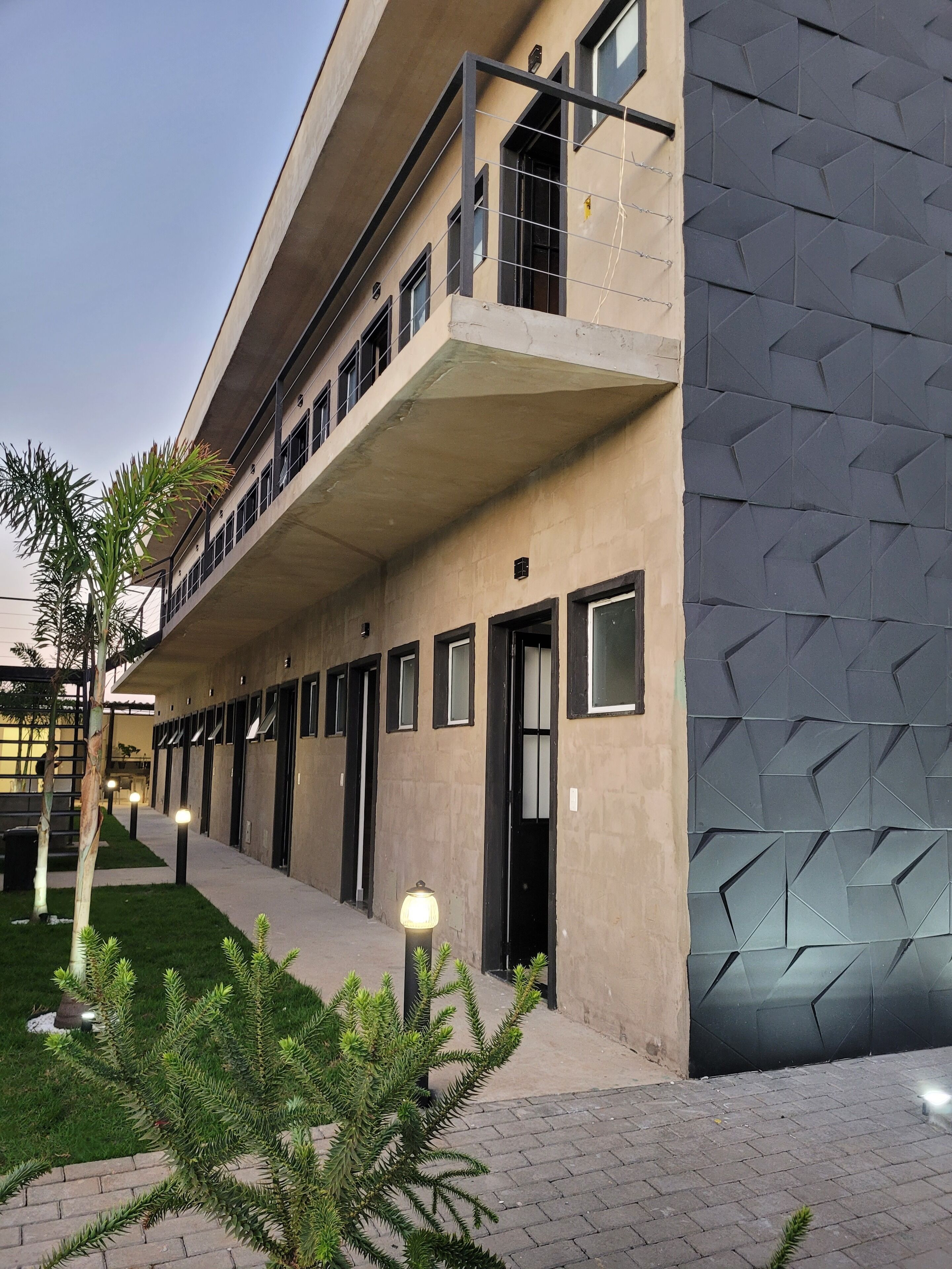 Un edificio moderno con fachada plana, varios ventanales y un balcón. Hay plantas en macetas y un paseo pavimentado.