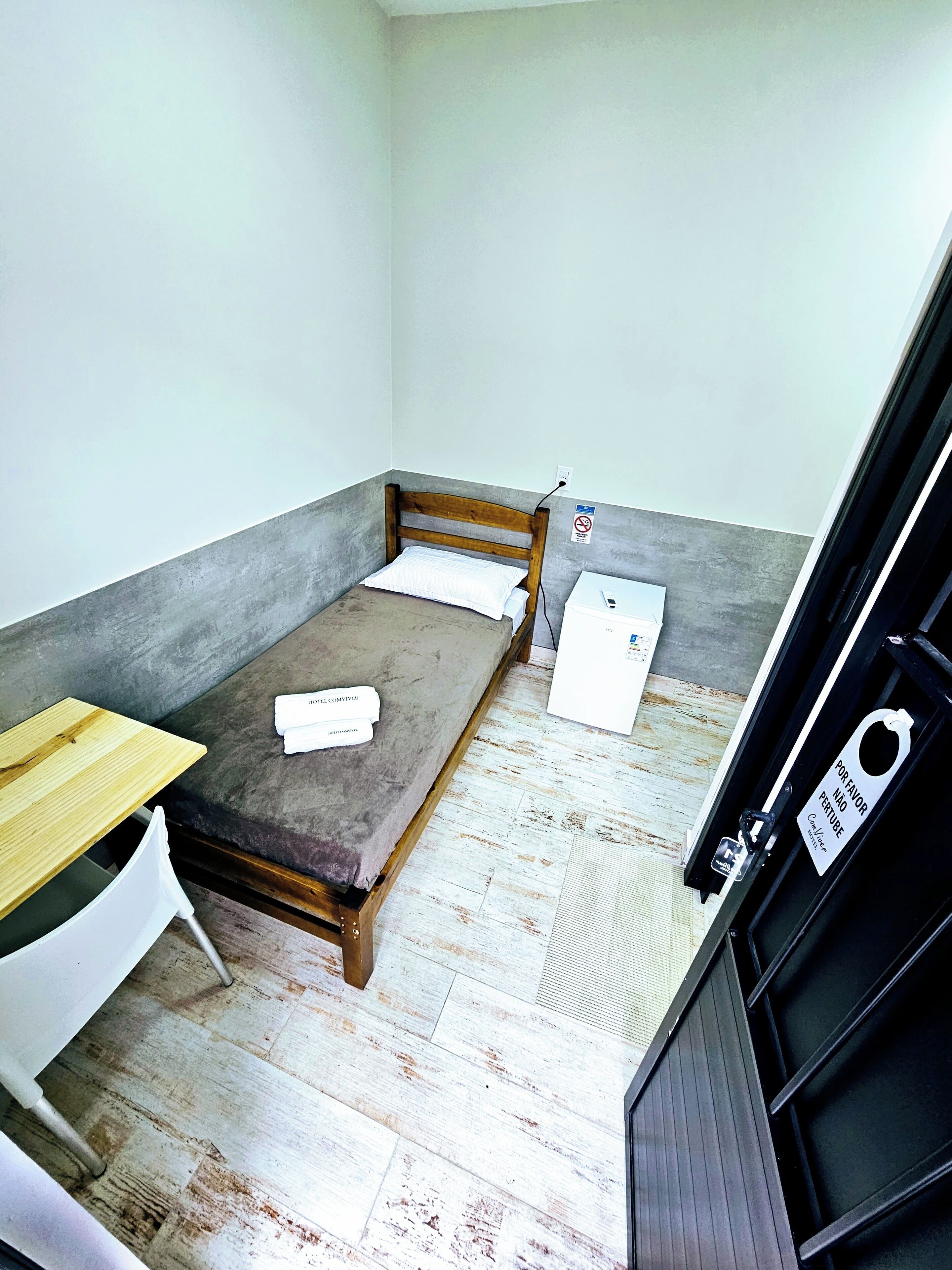Una habitación pequeña con cama individual, cabecera y mesa de madera, y una silla blanca. Hay una pizarra en la pared y un letrero con el símbolo de prohibido fumar.