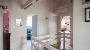Un pasillo con suelo de madera, paredes blancas y barandilla blanca. Se observan puertas que dan a otras habitaciones, y un tragaluz al fondo.