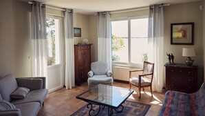 Una sala de estar con sofá, sillón, mesa de centro de vidrio y vistas al exterior a través de amplios ventanales.