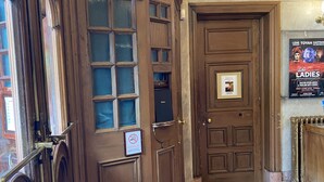 A wooden door labeled "STALLS" with a no-smoking sign, a poster for "Lene Toyah Saffron of Republic of the Ladies," and a patterned carpet leading to a room.