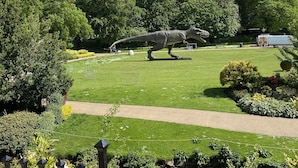 A dinosaur statue in a park with a pathway, trees, and a fence.