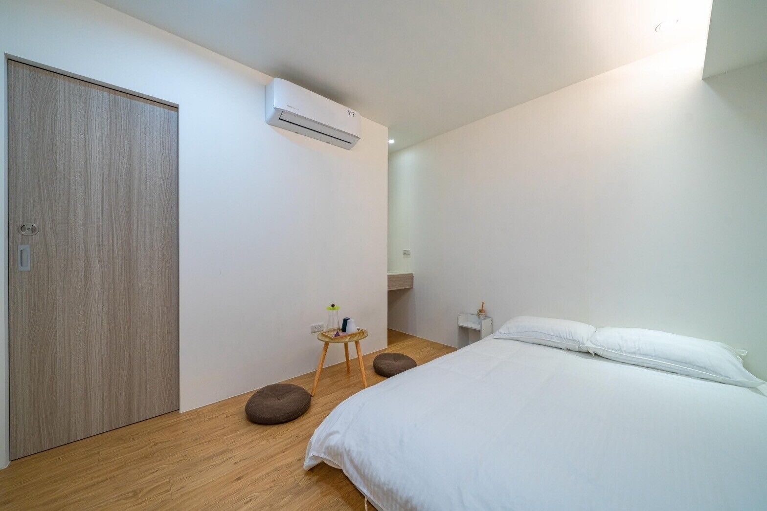 A bedroom with a bed, a wardrobe, an air conditioning unit, a small stool, and a bedside table with a lamp.