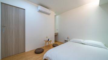 A bedroom with a bed, a wardrobe, an air conditioning unit, a small stool, and a bedside table with a lamp.