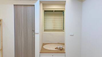 Una habitación pequeña con una puerta de madera, un ventanal con persianas, un banco de madera con una bandeja y un estante empotrado.