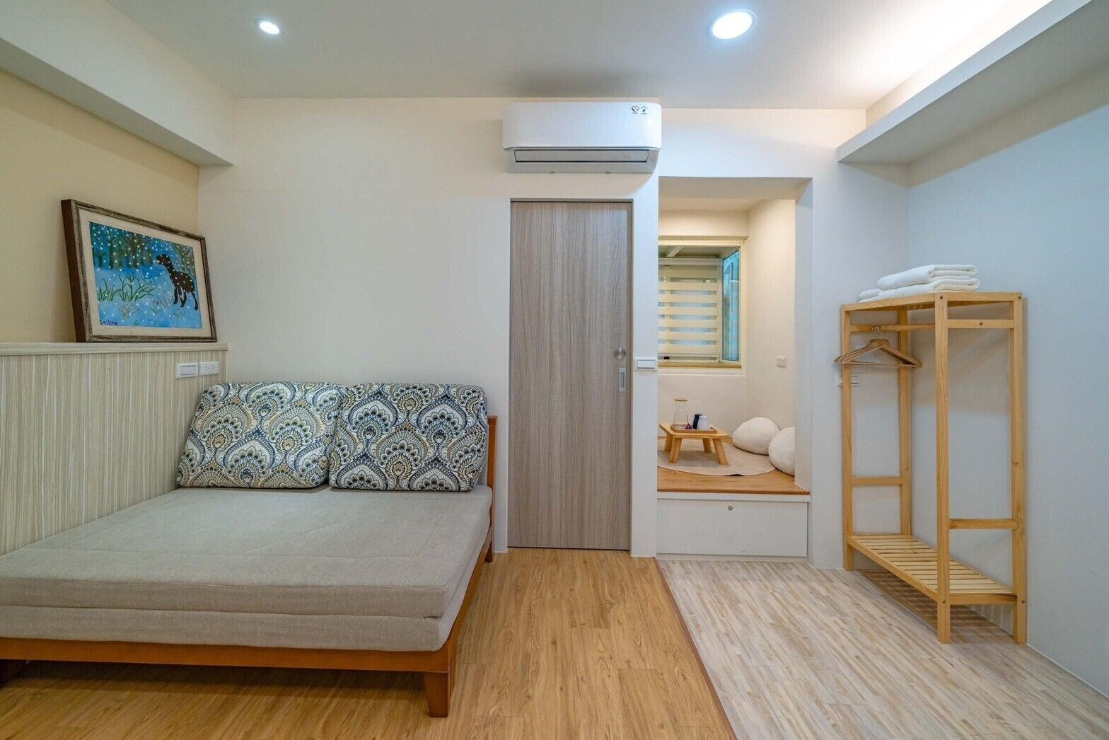 A bedroom with a bed, a wooden wardrobe, a small seating area with a window, and an air conditioning unit.