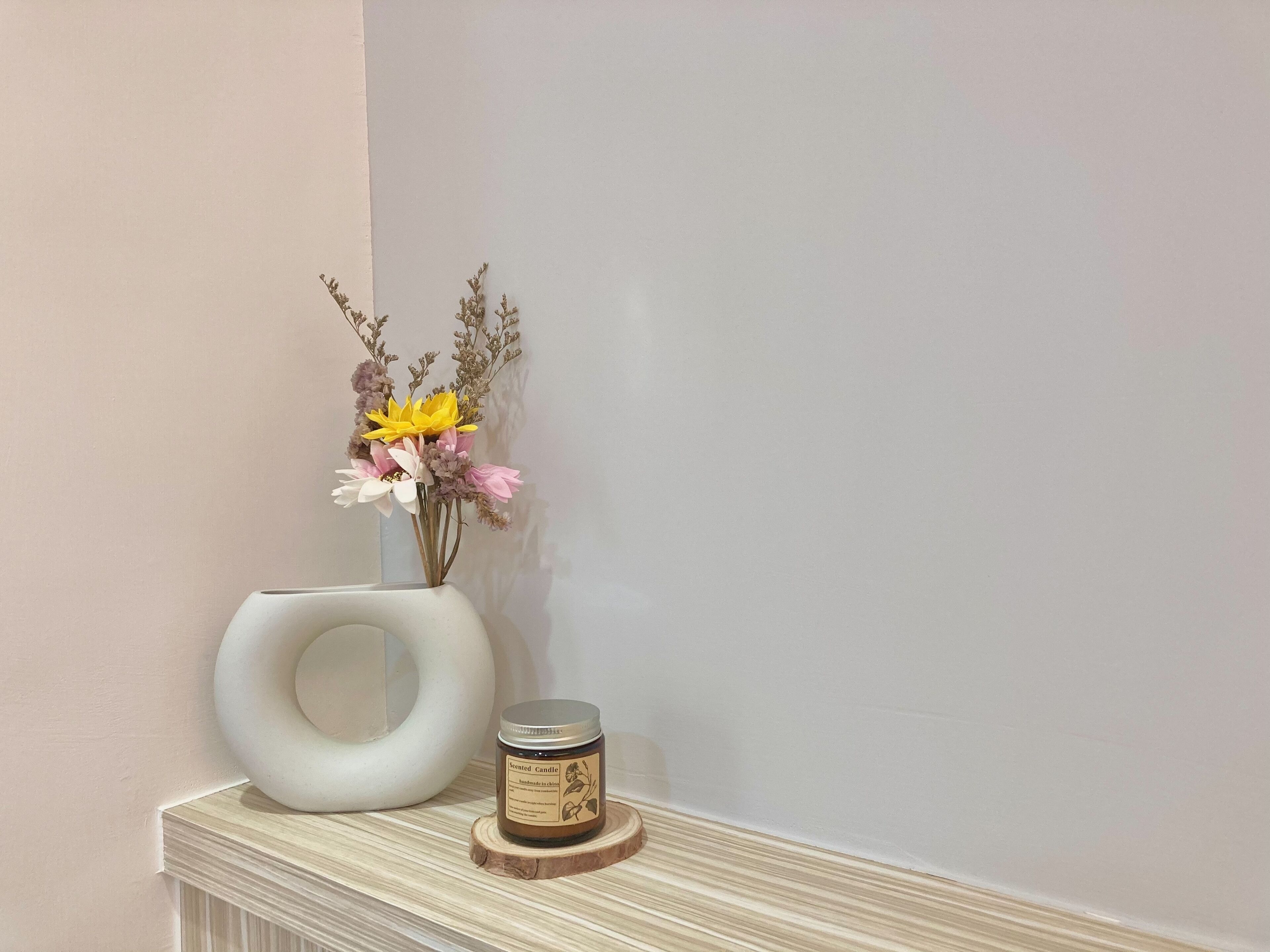 A white vase with flowers and a scented candle on a wooden coaster.