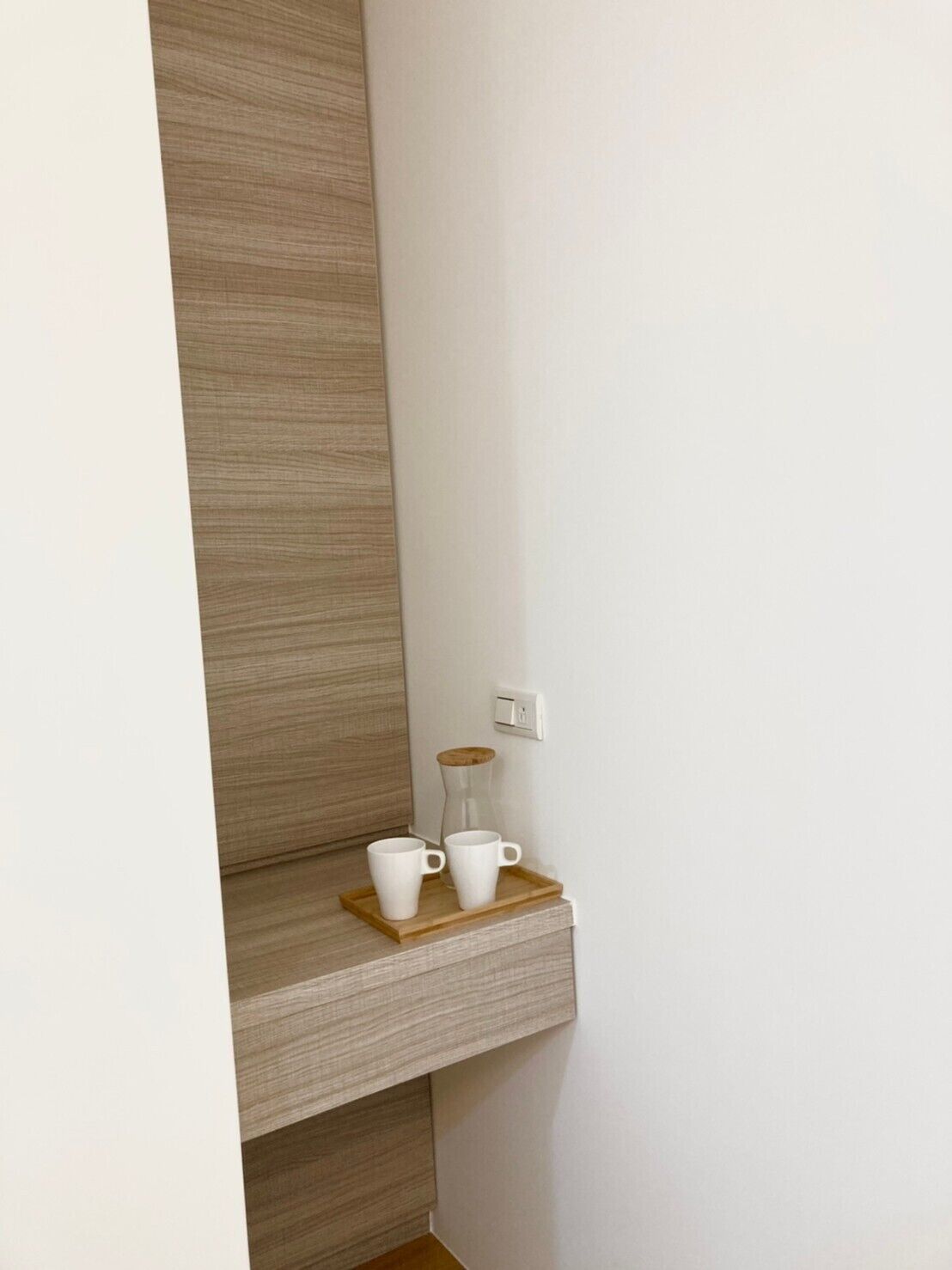 Un rincón de una habitación con una estantería de madera que sostiene dos tazas blancas y una jarra de vidrio.