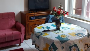 Un salon avec un canapé rouge, un téléviseur posé sur un meuble en bois, une table recouverte d’une nappe à motifs floraux et une fenêtre donnant sur un beau paysage.
