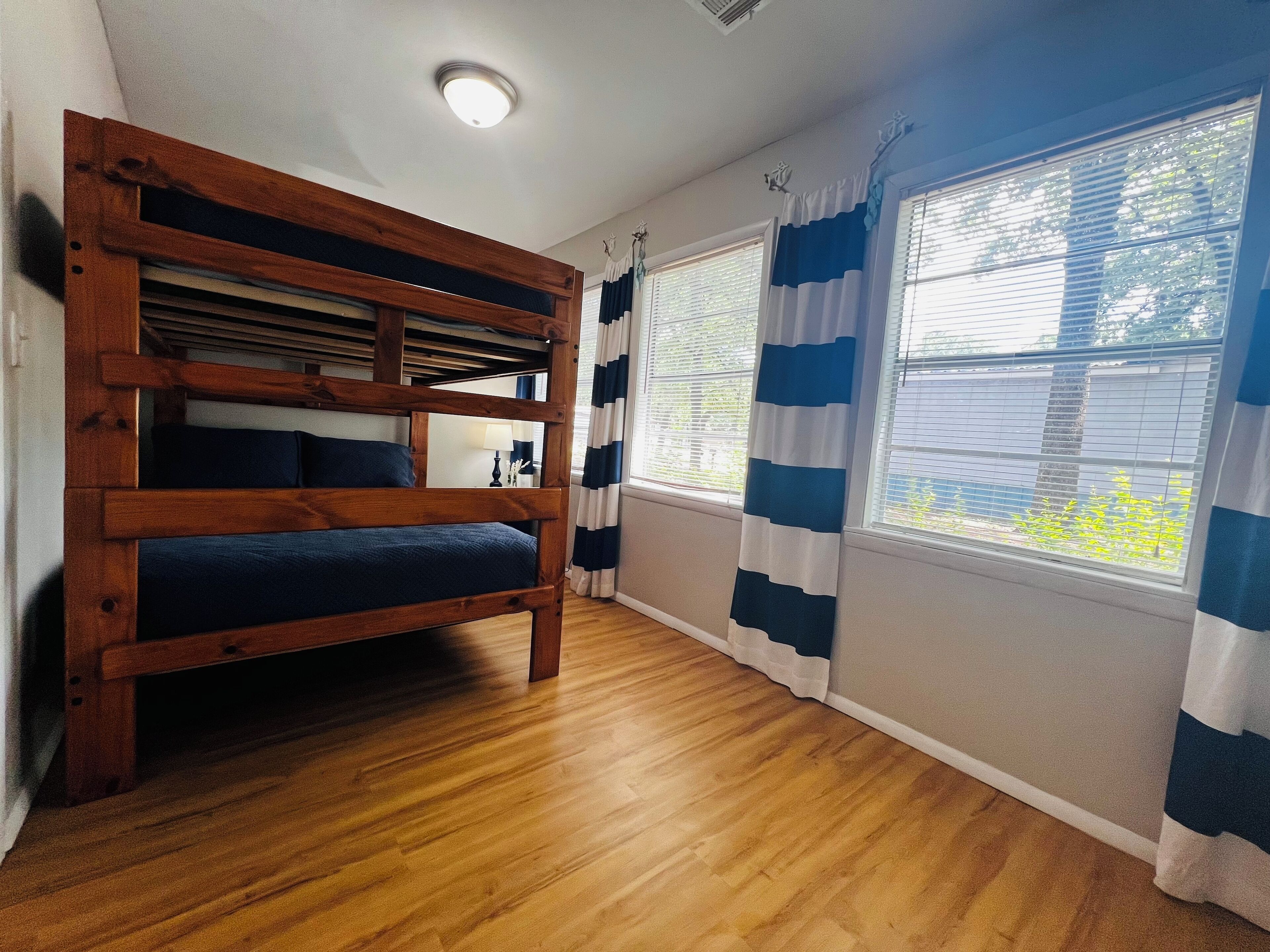 A room with a bunk bed, a window with blue and white curtains, and a wooden floor.