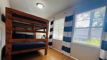 A room with a bunk bed, a window with blue and white curtains, and a wooden floor.