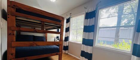 A room with a bunk bed, a window with blue and white curtains, and a wooden floor.