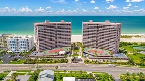 A high-rise hotel with a beach view, featuring multiple floors, a tennis court, and a parking area.